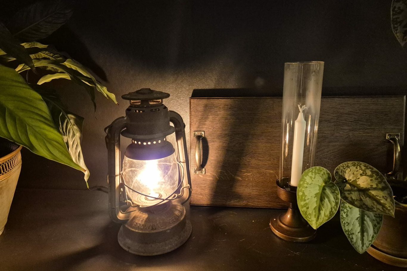 Designad lampa En gammal olje-lampa står på ett bord med gröna växter och ljus i bakgrunden.